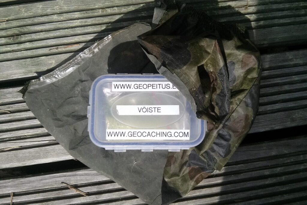 Geocache-Dose mit Beschriftung auf einem Tarnnetz auf Holzplanken