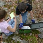Zwei Kinder öffnen eine gefundene Geocache-Kiste auf einem Felsen