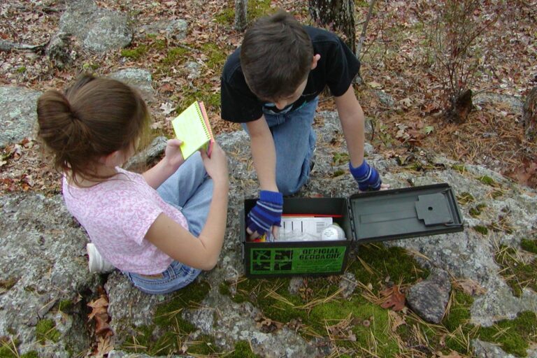 Zwei Kinder öffnen eine gefundene Geocache-Kiste auf einem Felsen