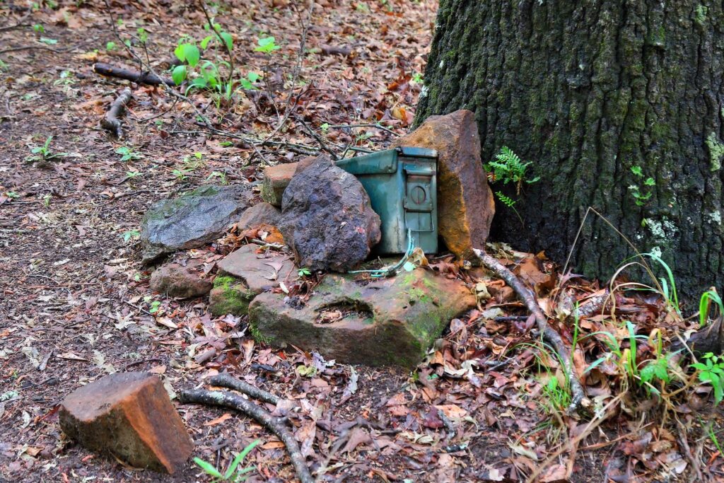 Grüne Munitionskiste als Geocache zwischen Steinen am Fuß eines Baumes