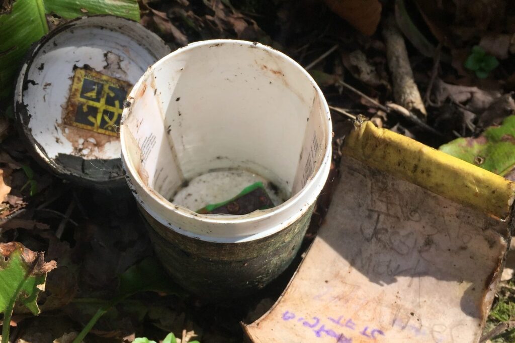 Geöffneter Geocache mit herausgezogenem, feuchtem Logbuch im Gras