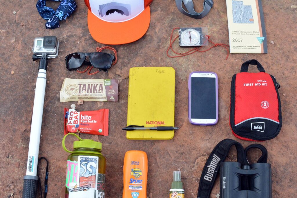 Wanderausrüstung ausgebreitet auf rotem Felsboden: Rucksack, Flasche, Fernglas, Smartphone und mehr