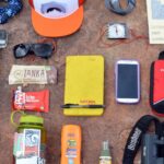Wanderausrüstung ausgebreitet auf rotem Felsboden: Rucksack, Flasche, Fernglas, Smartphone und mehr